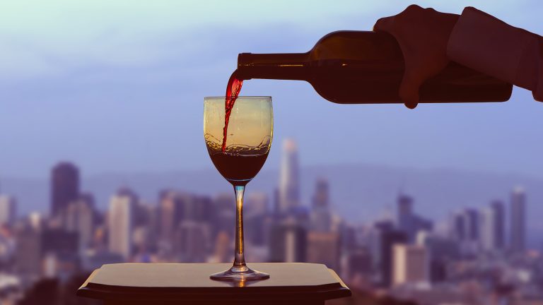 Female hand with bottle pours red wine into glasses on blurred San Francisco city background. Service on the roof of the restaurant